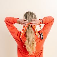 A woman with long blonde hair, seen from behind, wears an orange sweatshirt and ties her hair with a red Game Day Hair Scarf with Scrunchie by The Darling Effect—an ideal team spirit accessory. She also has a bracelet and a keychain with keys on her left wrist.
