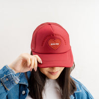 A person tilts down a Burgundy Game Day Trucker Hat by The Darling Effect, partially covering their face. They wear a blue denim jacket, adding subtle team spirit to their look.