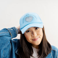 A woman with long dark hair wears The Darling Effect’s Light Blue Game Day Trucker Hat, a white top, and a blue denim jacket. She smiles softly, touching the back of her head while posing against a plain white background.