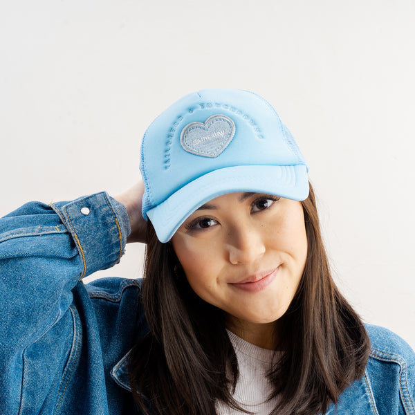 A woman with long dark hair wears The Darling Effect’s Light Blue Game Day Trucker Hat, a white top, and a blue denim jacket. She smiles softly, touching the back of her head while posing against a plain white background.