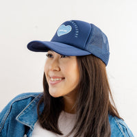 A woman with long dark hair smiles while wearing The Darling Effect's Navy Game Day Trucker Hat, a blue mesh cap with an adjustable back, paired with a denim jacket against a plain white background.