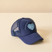 The Darling Effect Navy Game Day Trucker Hat features a blue mesh design, denim heart patch, light blue Sailing Day embroidery, and Tailoraries & Tour circular text. Adjustable mesh back ensures comfort. Shown on a light beige surface.