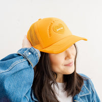 A person in The Darling Effect's Orange Game Day Trucker Hat and a blue denim jacket smiles, touching the cap and looking to the side, radiating team spirit.
