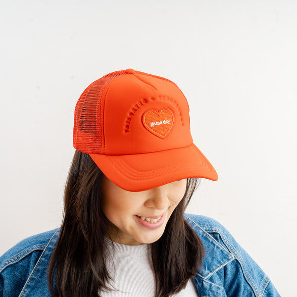 A woman with long dark hair smiles as she looks down, wearing The Darling Effect’s Red Game Day Trucker Hat with an embroidered heart and mesh adjustable back. She pairs it with a white shirt and blue denim jacket against a plain background.