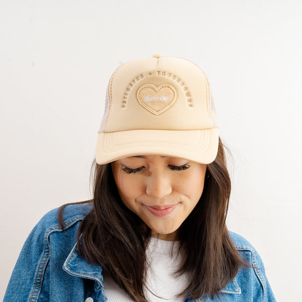 A woman wearing The Darling Effect’s Red Game Day Trucker Hat—featuring a heart-shaped patch on the front—and a light denim jacket looks down with a soft smile, her subtle team spirit shining through against a plain white background.