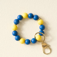 The Darling Effect’s Navy Yellow Game Day Hands-Free Wristlet features alternating blue and yellow beads, a gold ring, and a small charm engraved with “The Darling Effect,” arranged on a light cream background.