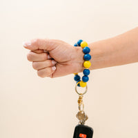 A hand wears The Darling Effect’s Navy Yellow Game Day Hands-Free Wristlet, looped around the wrist and holding keys with a car remote. The wristlet’s bold colors pop against a plain, light backdrop.