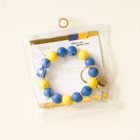 The Darling Effect Navy Yellow Game Day Hands-Free Wristlet features large blue and yellow beads, a gold keyring, and comes in a clear bag with a white “that’s so game day” card with gold accents—perfect for school pride.