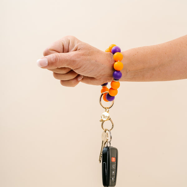 A hand wearing The Darling Effect Burgundy Gameday Hands-Free Keychain Wristlet—perfect for showing school spirit or matching your team’s colors—holds a keyring with keys and a car key fob against a plain light background.