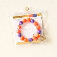 The Burgundy Gameday Hands-Free Keychain Wristlet by The Darling Effect, ideal for displaying school spirit or your favorite game day colors, rests in a clear plastic pouch against a light-colored wall.