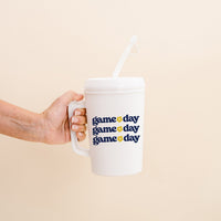 A hand holds The Darling Effect’s Navy Yellow Game Day 34 oz Mega Mug, featuring a handle, straw, and “game day” printed three times in bold blue and yellow on the front—perfect for tailgates and cheering on your team.