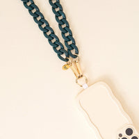 A close-up of The Darling Effect’s Dark Green Game Day Phone Chain with gold hardware, attached to a wavy-edged light phone case—stylishly displayed on a cream surface and perfect for your game day look.