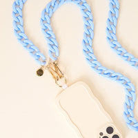The Darling Effect’s Light Blue Game Day Phone Chain, with gold clasps, is shown attached to a smartphone in a wavy beige case—perfect for game day style—displayed on a cream surface.