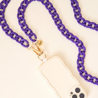 The Darling Effect's Purple Game Day Phone Chain features a chunky purple strap with gold hardware and a small charm, attached to a beige wavy-edged case—making it the perfect stylish accessory, shown on a light surface.