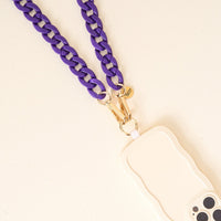 The Darling Effect’s Purple Game Day Phone Chain pairs a cream phone case with wavy edges, chunky purple strap, gold hardware, and charm—ideal for adding playful style to your look against a light background.