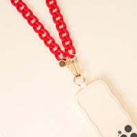 The Darling Effect’s Red Game Day Phone Chain, featuring gold hardware, is paired with a beige wavy-edged phone case for a bold look—perfect for game day and displayed on a light-colored surface.