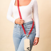 A person wearing a white long-sleeve top and blue jeans carries The Darling Effect’s Red Game Day Phone Chain with a beige scalloped phone case—an eye-catching crossbody accessory to elevate your game day style.