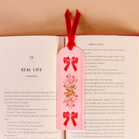 The Oh So Merry Acrylic Bookmark by The Darling Effect, featuring a vibrant floral design and a soft red velvet ribbon tassel, rests on an open book to mark the page titled REAL LIFE and HAPPY PLACE.