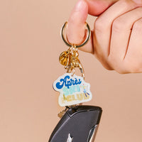 A hand holds a set of keys with The Darling Effect's Aprés Ski Club Acrylic Keychain, featuring blue, green, and yellow lettering on durable acrylic, plus a black car key and gold keyring.