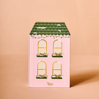 The Darling Effect's Classic Christmas Ornament Advent Calendar features a house-shaped pink box with four yellow-outlined windows, green flower boxes, a scalloped roof with string lights—perfect for displaying mini ornaments. Set against a peach background.
