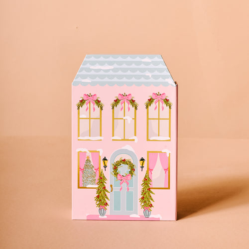 The Darling Effect’s Coquette Christmas Ornament Advent Calendar comes in a pink, house-shaped gift box decorated with wreaths, garlands, and bows—ideal for mini ornaments or beside Christmas trees. Shown against a peach background.