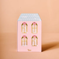 The Darling Effect’s Coquette Christmas Ornament Advent Calendar features a pink, house-shaped box with four front windows accented by pink bows and wreaths, a light blue scalloped roof—ideal for displaying mini ornaments or counting down to Christmas.