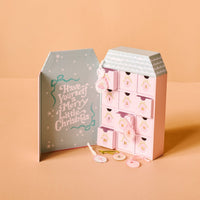 The Darling Effect’s Coquette Christmas Ornament Advent Calendar features pastel numbered drawers with animal designs, displayed next to a “Have Yourself a Merry Little Christmas” box and four sheep- and pig-shaped lollipops—ideal by an artificial tree.