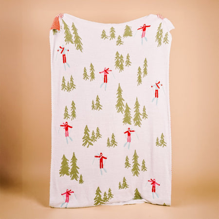 The Darling Effect Aprés Ski Cozy Dreams Luxe Blanket, featuring green trees and red skiers on white, is held by two hands at each top corner against a plain beige background.