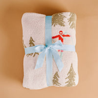 The Darling Effect Aprés Ski Cozy Dreams Luxe Blanket features a fuzzy white, ultra-soft polyester microfiber with a winter print of snow-covered trees and a skier, finished with a pale blue ribbon.