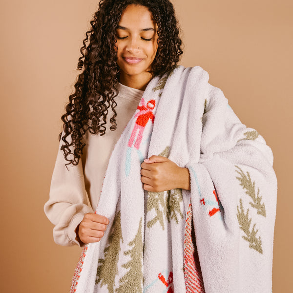 A young woman with curly hair, wearing a beige sweater, smiles gently while holding The Darling Effect’s Aprés Ski Cozy Dreams Luxe Blanket with colorful holiday and green tree patterns against a plain beige background.