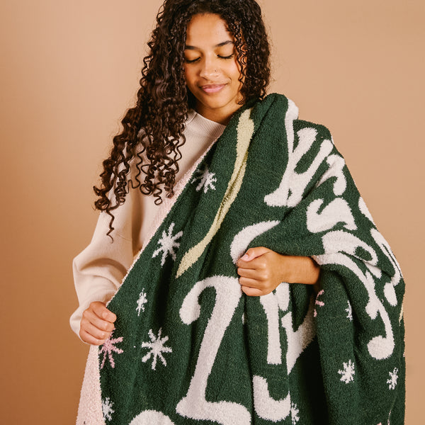 A woman with curly brown hair holds The Darling Effect's Merry Little Christmas Cozy Dreams Luxe Blanket, an ultra-soft microfiber throw with festive green and white snowflakes and a 2023 design.