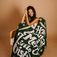 A smiling woman with curly hair sits on a chair, wrapped in The Darling Effect’s Merry Little Christmas Cozy Dreams Luxe Blanket, against a plain beige background.