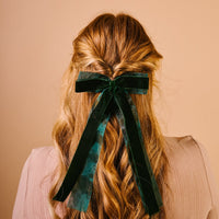 A person with long, wavy blonde hair styled half-up and adorned with The Darling Effect’s Green Velvet Luxe Layered Hair Bow faces away from the camera against a beige background. They wear a light-colored, ribbed shirt.