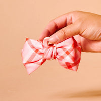 A hand holds The Darling Effect's Festive Plaid Mini Satin Bow Claw Clip, featuring pink and red satin plaid and a plastic claw, set against a beige background.