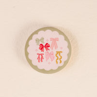 Store your Bow Affair Wood Enamel Coasters from The Darling Effect in style with this round tin, featuring a scalloped white label adorned with six colorful illustrated bows arranged in a circle on a light background.