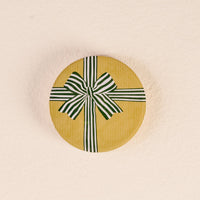The Darling Effect’s Holiday Gift Bow Green Wood Enamel Coaster comes in a round yellow box with subtle stripes and a bold green-and-white bow, perfect for gifting, set against a textured light beige background.