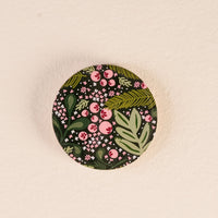 A round Jolly Sprig Wood Enamel Coaster from The Darling Effect, featuring a green, pink, and white floral pattern, rests on a textured, light-colored surface—making it an ideal decorative accent for your table. Both stylish and functional, this coaster brings charm and protection to your home décor.