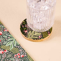 A glass filled with ice rests on the Jolly Sprig Wood Enamel Coaster by The Darling Effect, which showcases a round design adorned with leafy and red berry patterns. Beside it lies a green fabric or napkin featuring a matching motif, all displayed on a light beige surface. Perfect for entertaining, these coasters from The Darling Effect combine durability and festive style for your tabletop setting.