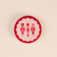 The Darling Effect’s Nutcrackers Wood Enamel Coaster showcases three pink and red nutcrackers on a white background with a red border, adding festive charm to your holiday gatherings.