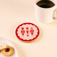 A round cookie with three nutcracker soldiers sits beside a white mug of black coffee on The Darling Effect's Nutcrackers Wood Enamel Coaster, with more cookies and a wafer on a plate, all on a light surface.