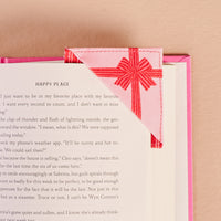 A close-up of an open book with The Darling Effect’s Holiday Gift Bow Red Corner Keeper Fabric Bookmark—featuring a pink and white plaid pattern with a red bow—placed on a light pink surface, making it the perfect gift for any book lover.