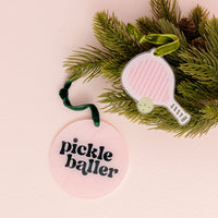 Two festive decorations hang from pine branches: one is The Darling Effect’s Holiday Tree Ornament - Pickleballer, a round pink acrylic ornament featuring “pickle baller” in bold black text, and the other is shaped like a pink-striped pickleball paddle—perfect touches for the holiday season.