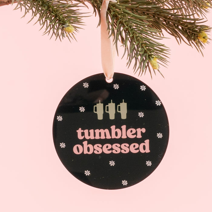 The Darling Effect’s Holiday Tree Ornament - Tumbler Obsessed is a round black acrylic ornament featuring the phrase “tumbler obsessed,” three tumbler icons, and delicate snowflakes. Hanging from a pine branch against a pink background, it’s the perfect festive addition to your holiday décor.