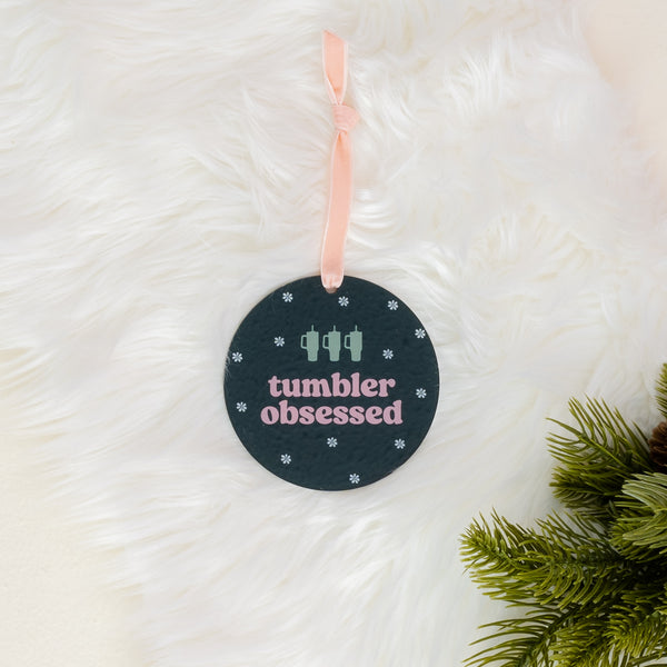 The Darling Effect’s Holiday Tree Ornament – Tumbler Obsessed features a round acrylic design with “tumbler obsessed” in pastel letters, two cup icons, and small snowflakes. Hanging from a pink ribbon on a white furry background with green pine branches, this ornament makes the perfect festive decoration for the holiday season.