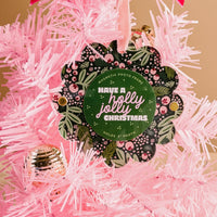 A close-up of The Darling Effect’s Jolly Sprig Scalloped Round Picture Frame Ornament, featuring a green floral design and “Have a holly jolly Christmas” text, displayed on a pink artificial tree next to a shiny gold ornament. Holds a 2" photo.