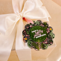A gold gift box tied with a cream ribbon features The Darling Effect's Jolly Sprig Scalloped Round Picture Frame Ornament, which says "Have a holly jolly Christmas" and holds a 2" photo.