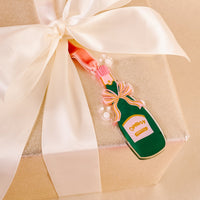 A gold gift box with a large ivory satin ribbon is topped with The Darling Effect's Champagne Bottle Holiday Tree Ornament, featuring a green bottle, pink bow, bubbles, and yellow label—perfect for the festive season.