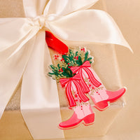 The Darling Effect’s Festive Cowgirl Holiday Tree Ornament comes in a gold gift box with a white satin ribbon, topped with a tag featuring pink cowboy boots filled with greenery, red berries, and pink bows for the holidays.