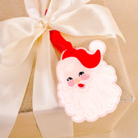 A gift wrapped in gold paper with a white satin ribbon, topped with a gift tag featuring a cartoon Santa Claus face wearing a red hat—perfect for adding holiday charm and pairing beautifully with festive décor or The Darling Effect’s Santa Shape Tree Ornament.