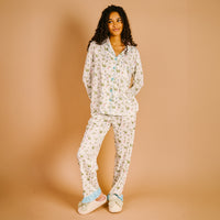 A woman wears The Darling Effect Aprés Ski Sweet Dreams Pajama Pants Set, featuring a white long-sleeve top and matching pants with a winter forest pattern, light blue trim, and ruffled cuffs.